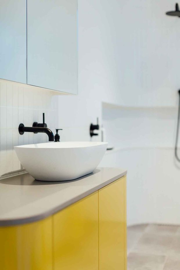 Bright contemporary bathroom featuring a white vessel basin on a pale stone benchtop, vivid yellow built-in vanity cabinetry, matte black wall-mounted tapware, white tiled wall surfaces, and a large mirror. The composition focuses on the vanity area, with a shower fixture partially visible to the right.