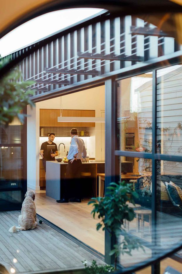 A warmly lit contemporary home interior viewed through large black-framed glazing from a timber deck. The image focuses on a compact kitchen with a central island, timber cabinetry, pale benchtops, pendant lighting and two people standing at the island. A small dog sits on the deck in the foreground, while reflections and views through the glazing reveal an adjoining living area and a multi-level courtyard-like architectural setting.