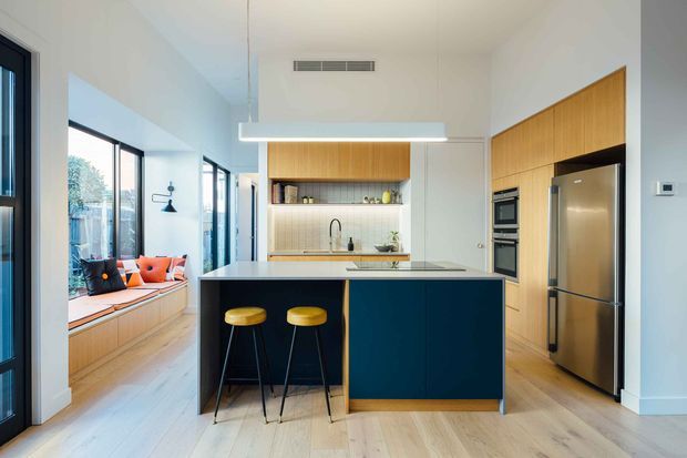 Bright contemporary open-plan kitchen with a large dark blue island, integrated timber cabinetry, stainless-steel appliances, and two bar stools. A built-in window seat with colourful cushions is visible along the glazed side of the room. The space has pale timber flooring, white walls, a tiled backsplash, and a linear suspended light.