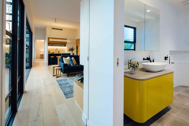 A bright contemporary interior showing a compact bathroom in the foreground, with a vivid mustard-yellow floating vanity, pale stone benchtop, white vessel basin, wall-mounted black tapware, and large white wall tiles. Through the open doorway is an open-plan living and kitchen area with pale timber flooring, dark-framed glazing, a navy sofa, and built-in kitchen cabinetry. The spaces are connected by a clean white hallway and strong indoor-outdoor glazing.