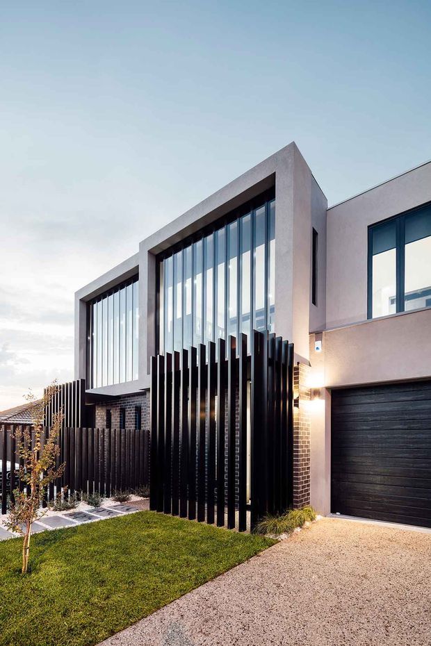 Contemporary multi-storey residential facade viewed from the driveway, featuring pale rendered upper walls, dark brickwork, dramatic floor-to-ceiling vertical glazing, black vertical metal privacy screens, and a dark garage door. A small landscaped front yard and concrete driveway complete the street-facing composition.