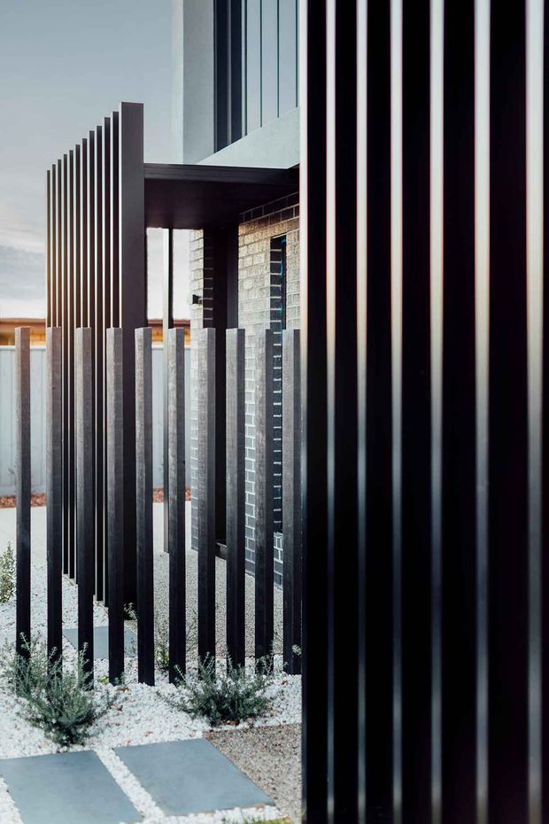 A close architectural view of a contemporary building facade and entry screen. Repeated tall, narrow dark metal fins create a strong vertical rhythm in the foreground and along the left side, partially concealing a recessed entrance beneath a flat canopy. Grey masonry walls, dark-framed glazing, pale gravel landscaping, and large rectangular stepping pavers are visible. The composition is stark, geometric, and highly textural, with the black metal screening dominating the image.
