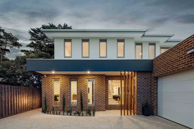 Contemporary two-storey residential home viewed from the driveway, featuring a red-brown brick ground floor, smooth pale upper-level walls, narrow vertical windows, a deep dark blue-grey projecting balcony or awning, and a low-pitched roof with broad eaves. A timber screen marks the illuminated entry beside an attached garage. The foreground includes a textured paved driveway and timber boundary fencing.