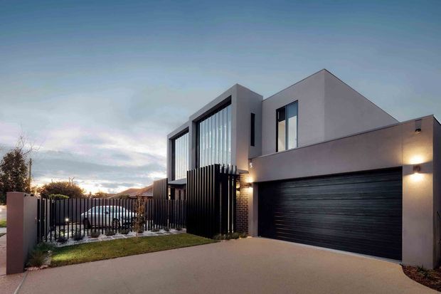A contemporary multi-storey residence viewed from the street at dusk. The facade combines smooth pale rendered walls, dark brickwork, extensive vertical glazing, black vertical screening and a large dark sectional garage door. A paved driveway, front fence and small landscaped garden sit in the foreground, with warm exterior wall lighting highlighting the entry and garage.