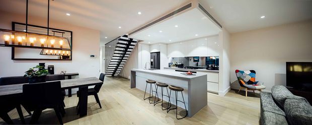 Open-plan contemporary interior combining a dining area, kitchen and living room. The space features pale timber flooring, white walls and cabinetry, a large island with integrated seating, black-framed open-riser stairs, recessed lighting, and a linear black metal pendant above the dining table. A grey sofa and colourful accent chair furnish the living area.