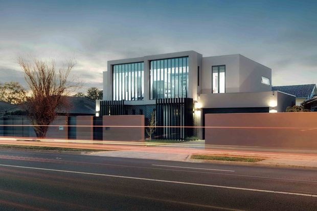 Street-facing exterior view of a contemporary two-storey residence at dusk. The house has a boxy flat-roofed form, smooth light-grey rendered walls, extensive floor-to-ceiling glazing with vertical fins, dark metal screening, a garage, and a low rendered front boundary wall. Motion-blurred vehicle light trails run across the foreground street.