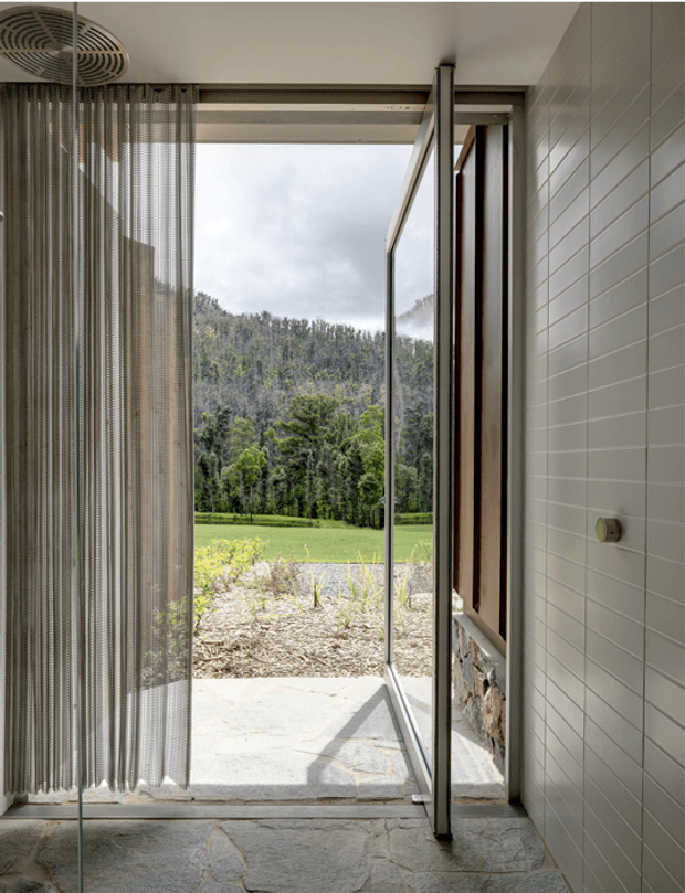 A bright, narrow interior threshold opening directly onto a landscaped rural garden. The space features pale irregular stone flooring, white horizontal wall tiles, slim dark-framed full-height glazing and an open glass door. A vertical chain curtain hangs beside the opening, while a covered outdoor terrace and green field with wooded hills are visible beyond. The image emphasizes indoor-outdoor connection, natural light and a restrained contemporary material palette.