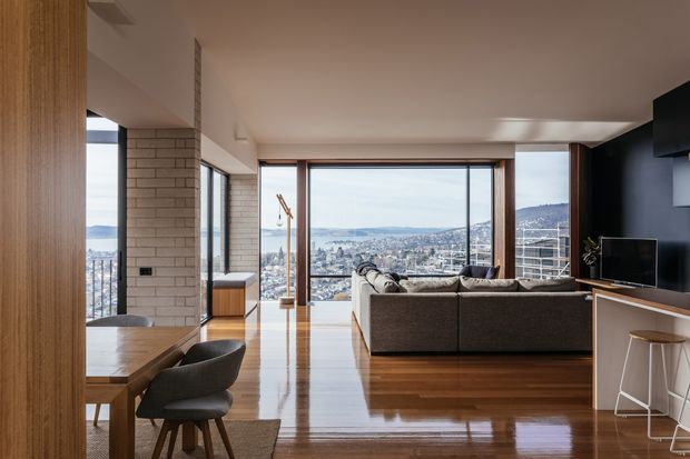 A contemporary open-plan living and dining area with expansive floor-to-ceiling glazing overlooking a dense hillside cityscape. The room features polished timber flooring, warm timber wall panelling and cabinetry, a pale brick column, a large sectional sofa, a dark feature wall with television, and a compact breakfast bar. Natural daylight and the elevated urban outlook create a refined, spacious interior.