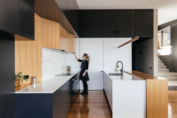 A contemporary galley-style kitchen with dark navy-black cabinetry, pale timber wall panelling, white benchtops and a glossy dark timber floor. A large white island with integrated sink and curved black tap dominates the foreground, while a black tiled feature wall and open stairway are visible at right. The space has clean lines, integrated appliances, a high ceiling and a restrained monochromatic palette with warm timber accents.