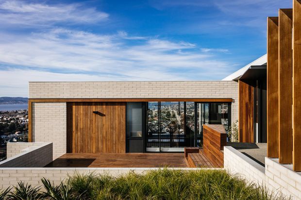 Exterior view of a contemporary multi-level residence on a hillside, featuring pale brickwork, warm vertical timber cladding, timber-framed glazing, a broad flat roofline, and a rooftop terrace with built-in planters. A coastal urban landscape is visible beyond the house.