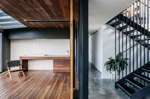 Split view of a contemporary interior showing a timber-lined room on the left and a minimalist hallway with a black open-riser staircase on the right. The left space features warm timber flooring and ceiling, a white brick feature wall, and built-in timber cabinetry with a dark benchtop. The right side has polished grey concrete flooring, white walls, dark metal stair elements, and a potted plant.