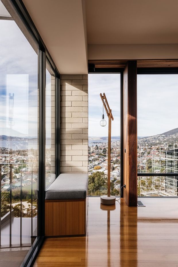 A bright contemporary interior corner with expansive floor-to-ceiling glazing overlooking a hilly urban landscape. The room features warm polished timber flooring, exposed light masonry walls, dark-framed windows, a built-in timber bench with a grey cushion, and a sculptural timber floor lamp. Strong vertical structural framing and panoramic views define the space.