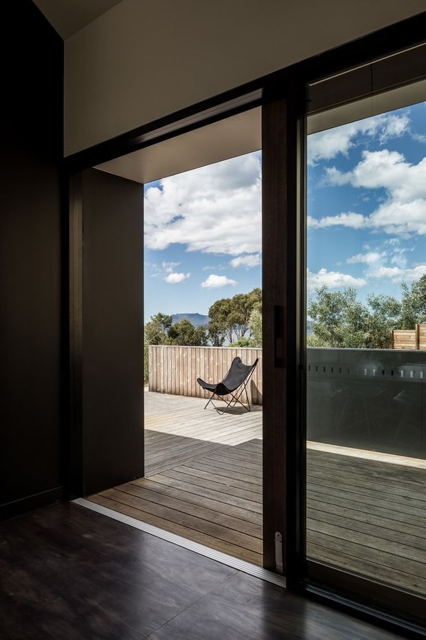 A dark, minimalist interior opens onto a timber deck through large floor-to-ceiling glazed sliding doors. The deck includes a black sling chair and overlooks trees, a distant hill, and bright blue sky. Deep charcoal or black interior surfaces and timber decking create strong contrast with the outdoor landscape.
