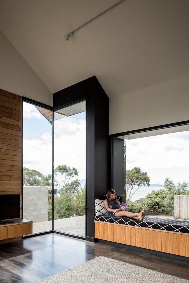 A contemporary living room with expansive black-framed corner glazing overlooking trees and water. The room features a built-in timber window seat with black-and-white cushions, dark timber flooring, a timber-lined feature wall, a low timber media console, and a large pale rug. A person is seated on the window bench using a tablet. The space is bright, open, and connected to the landscape.