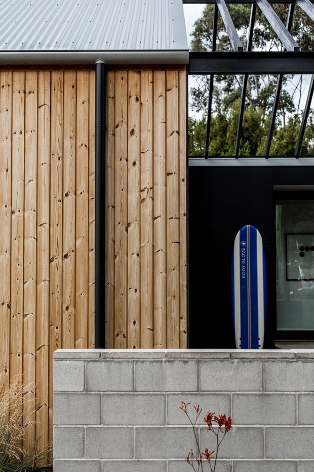 Close-up exterior view of a contemporary building combining vertical natural timber cladding, a light corrugated metal roof, black-framed glazing and dark structural beams. A grey concrete block privacy wall occupies the foreground, with a visible outdoor shower or entry area and a blue bodyboard leaning against the facade. Trees are visible through the glazing.