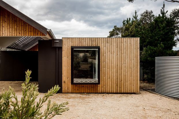 Exterior view of a compact timber-clad building or extension with a steeply pitched roof section, dark metal detailing, and a central reflective window. The structure sits in a gravelled landscaped setting with mature trees and a large corrugated cylindrical water tank visible at right. The timber cladding and roof forms create a warm, contemporary rural character.