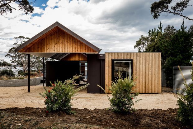Exterior view of a compact contemporary timber-clad building with a prominent gabled roof and deep covered entry or outdoor living area. The facade combines warm vertical timber boards, dark wall panels, exposed timber soffit lining, and large glazed openings. It sits in a landscaped, semi-rural setting with gravel around the structure and newly planted shrubs.