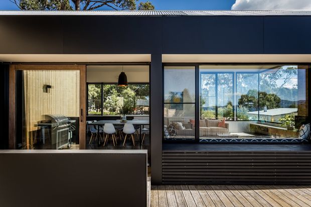 Contemporary single-storey residential facade with dark horizontal cladding and broad glazed openings. The view shows a dining area and living room through the windows, with a timber deck in the foreground and reflected landscaping and sky in the glazing. A dark projecting roof or canopy creates a strong horizontal composition.