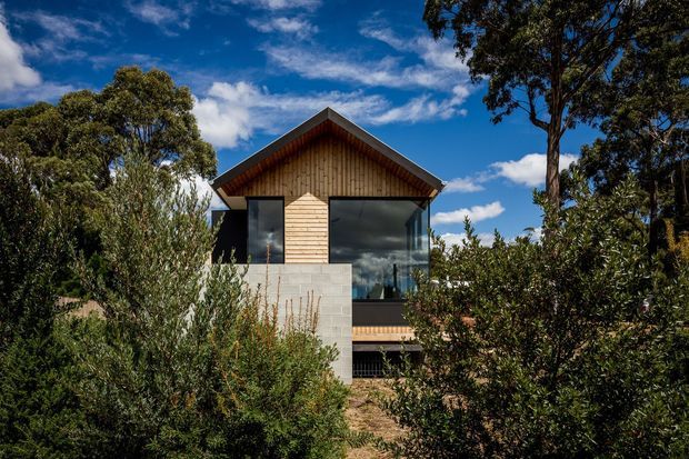 Exterior view of a compact contemporary home set among dense native vegetation. The building has a prominent timber-clad gabled upper facade, dark-framed glazing, and a pale masonry lower section, partially screened by landscaping.
