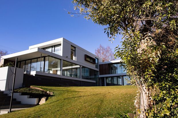 Exterior photograph of a contemporary multi-level residence on a sloping landscaped site. The house features crisp white rendered volumes, extensive floor-to-ceiling glazing, dark horizontal cladding, projecting flat-roofed forms, and connected pavilion-like wings. Mature trees and lawn frame the foreground.