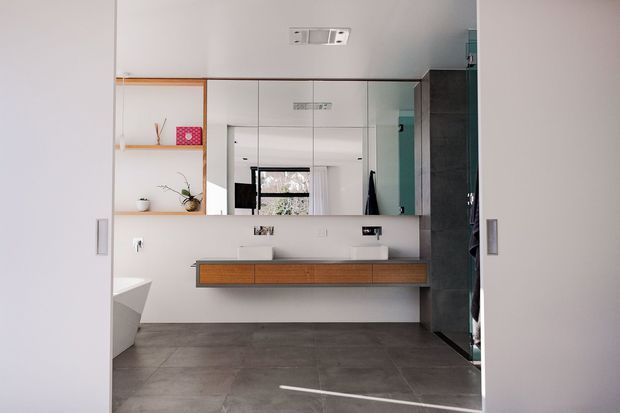 Contemporary bathroom viewed through partially open doors, featuring a long floating timber vanity with integrated basins, large wall-to-wall mirror, grey tiled floor, white walls, open shelving, and a partially visible freestanding bathtub. The room has a clean, minimalist fit-out with natural timber accents and abundant reflected daylight.