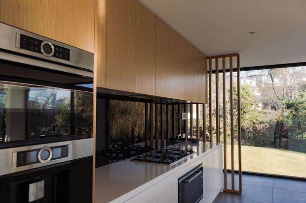 A contemporary kitchen with extensive warm timber overhead cabinetry, integrated stainless-steel ovens, a glossy black reflective splashback, white stone island benchtop, and a gas cooktop. Full-height glazing and a vertical timber screen open the kitchen to a landscaped garden, while dark flooring and minimal detailing create a refined, light-filled space.
