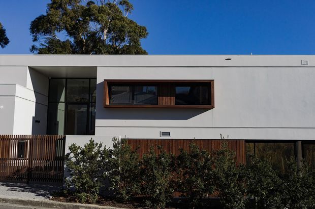 Street-facing view of a contemporary single-storey flat-roof house with a crisp white rendered facade, deep-set dark glazing, warm timber window framing and vertical timber screening. Low landscaping partially obscures the lower facade, while a timber-slatted entry fence and recessed entry create privacy.