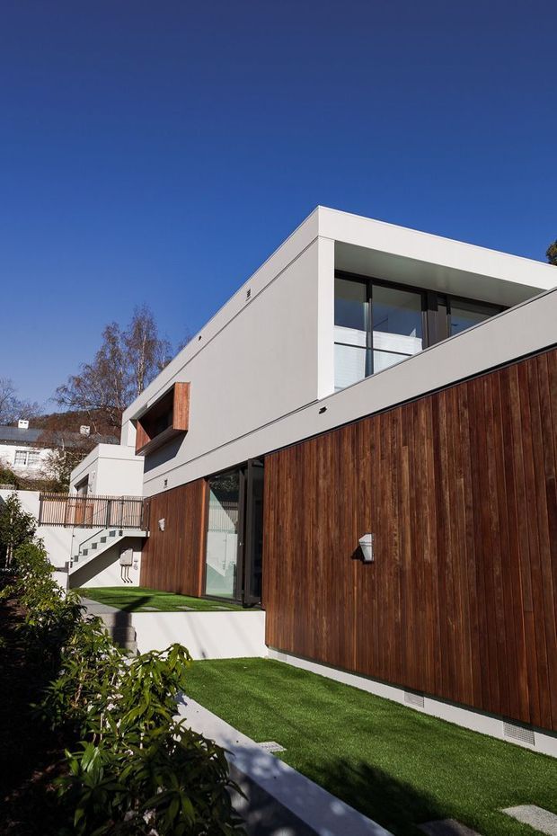 Exterior view of a contemporary multi-level residence with crisp white rendered volumes, dark-framed glazing, and extensive vertical timber-clad walls. The home sits on a landscaped sloping site with stepped paths, a raised terrace, balcony, and external stairway under a clear blue sky.