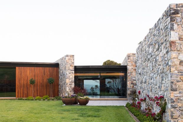 Single-storey contemporary residence with a low, flat roof and extensive glazing, framed by rugged natural stone walls and stone piers. A warm timber-clad entry volume, dark metal framing, landscaped lawn, and sculptural planters create a refined, minimalist courtyard-like frontage.