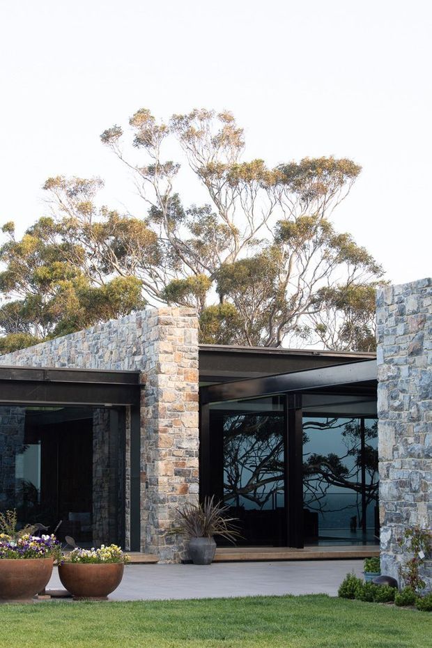 Exterior view of a contemporary low-profile building with rough-faced natural stone walls, dark steel roof framing, and expansive black-framed glazing. The entrance and covered terrace open onto a manicured lawn with large planter pots, while mature eucalyptus trees are visible behind and reflected in the glass.