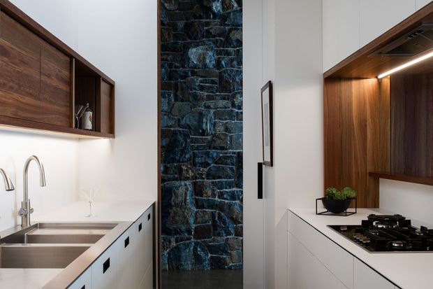 A compact, highly resolved contemporary kitchen viewed toward a dark natural-stone feature wall. The space includes white lower cabinetry, dark walnut-toned overhead and tall joinery, stainless-steel double sinks and tapware, a gas cooktop, integrated task lighting, and a small plant on the benchtop. The palette combines white, dark timber, black and deep blue-grey stone, creating a refined, high-contrast interior.
