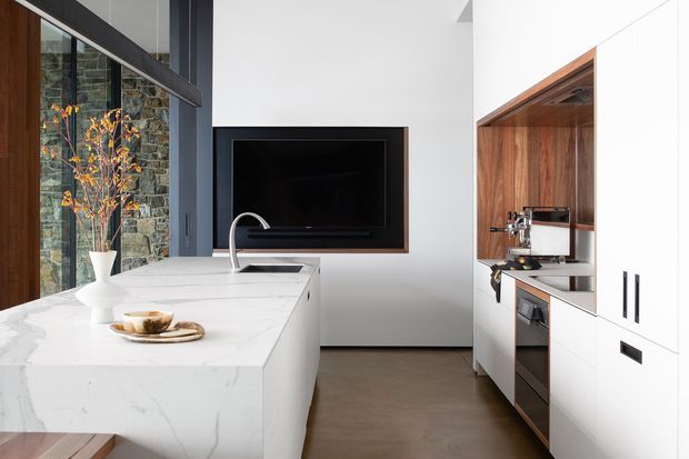 A bright contemporary kitchen with a large white marble island, integrated sink and curved faucet, white cabinetry, and a warm timber-lined coffee or appliance nook. A dark stone feature wall, extensive glazing, and a wall-mounted television create contrast, while the concrete-toned floor and black accents reinforce the modern design.
