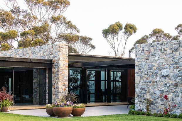 Low-profile contemporary home set within a landscaped garden, featuring extensive floor-to-ceiling dark-framed glazing, flat roof forms, and substantial blue-grey natural stone masonry walls. Native trees and sculptural planters surround the building.