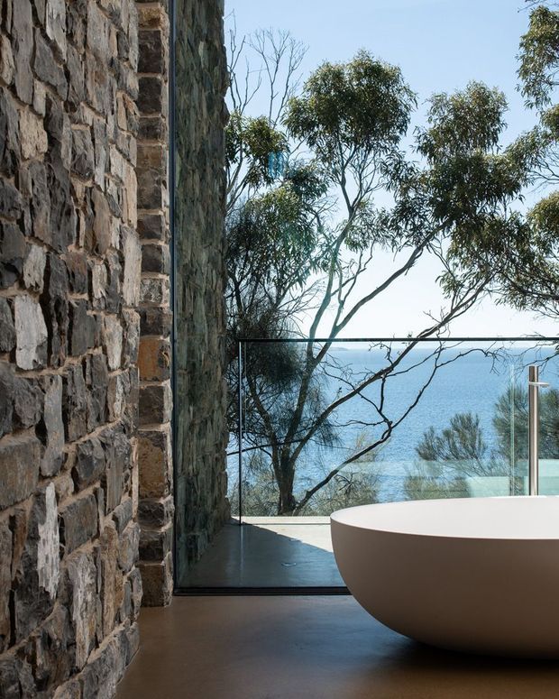 A dramatic bathroom interior with a freestanding white oval bathtub beside a full-height glazed opening overlooking blue water and coastal vegetation. A rough natural stone feature wall contrasts with the smooth dark floor and frameless glass balustrade, creating a refined, nature-connected setting.
