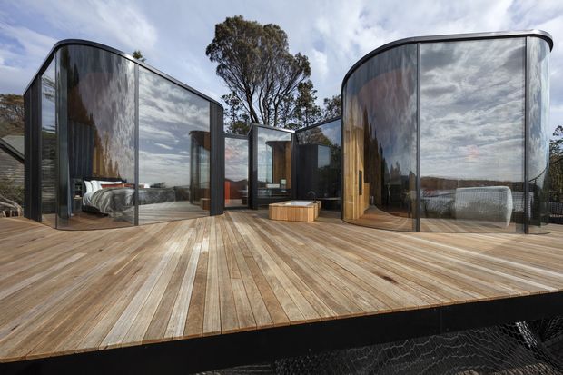 Exterior view of a contemporary pod-style dwelling or retreat on a raised timber deck. Several rounded, dark-framed glazed volumes feature highly reflective curved glass, with bedrooms and living spaces visible inside. The structure is surrounded by trees and sits above a dark supporting base, creating a sculptural, lightweight architectural composition.