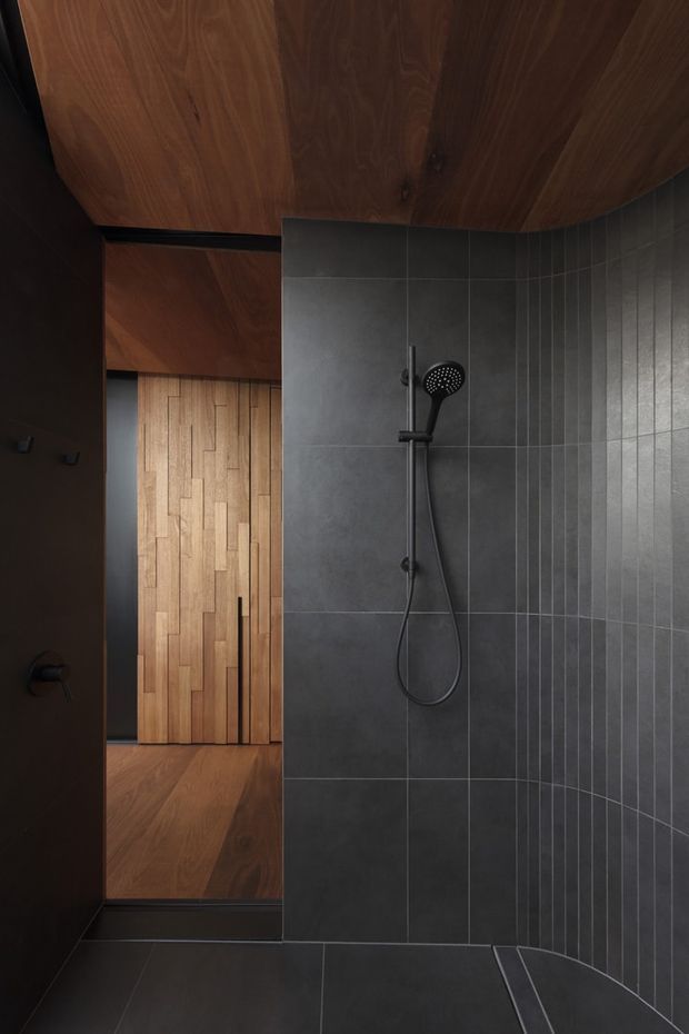 A dark, minimalist walk-in shower with charcoal grey wall and floor tiles, matte black shower fittings, and warm timber wall and ceiling surfaces. A timber-lined passage or doorway is visible beyond the shower, creating a strong contrast between the cool tiled wet area and warm wood interior.