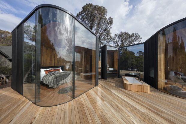 Exterior view of a contemporary multi-pavilion dwelling with dramatic curved floor-to-ceiling glazing, dark cladding, warm timber interiors, and a broad timber deck. A bedroom is visible through the glass, with additional living spaces and outdoor seating arranged around the deck. The curved glazed forms and surrounding trees create a strong connection to the landscape.