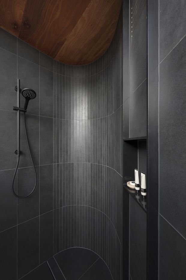 A compact, dark contemporary shower enclosure with charcoal large-format wall tiles and a narrow vertical-ribbed feature tile panel with curved line detailing. A black handheld shower is mounted on the left, while a recessed black shelf on the right holds toiletries. The ceiling includes a curved, warm-toned timber-lined section, creating contrast against the predominantly black and grey finishes.
