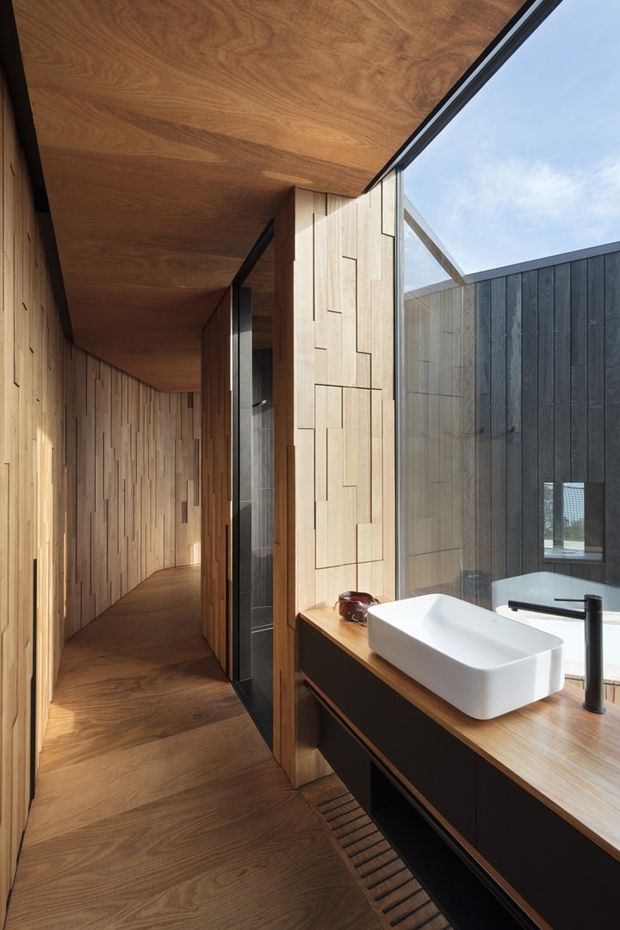 A refined contemporary bathroom with a white vessel basin, matte black tapware, and dark built-in vanity topped with warm timber. The room is wrapped in detailed natural timber wall and ceiling panelling, with matching timber flooring and a large glazed opening that brings in daylight and views of an adjacent dark-clad exterior wall. A narrow timber-lined corridor extends beyond the vanity.