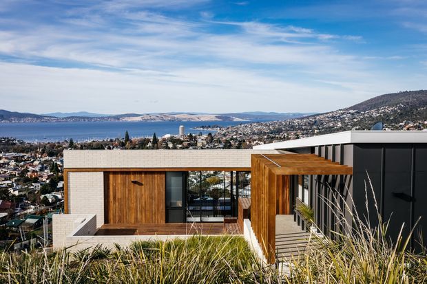 Modern multi-level residential building on a hillside overlooking a bay and dense coastal settlement. The architecture features crisp rectilinear volumes, pale masonry walls, extensive glazing, warm timber-clad sections, and a dark metal-clad upper volume. Native grasses occupy the foreground roof or landscaped terrace.