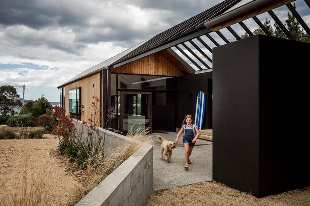 Exterior view of a contemporary coastal residence with timber-clad walls, a steep pitched dark roof, and a dramatic glazed entry canopy with exposed structural beams. A black rendered volume frames the entrance, while a landscaped sandy garden and low concrete-block retaining wall lead toward the home. The image also shows a child and dog near the entry, with glimpses of the sea beyond.