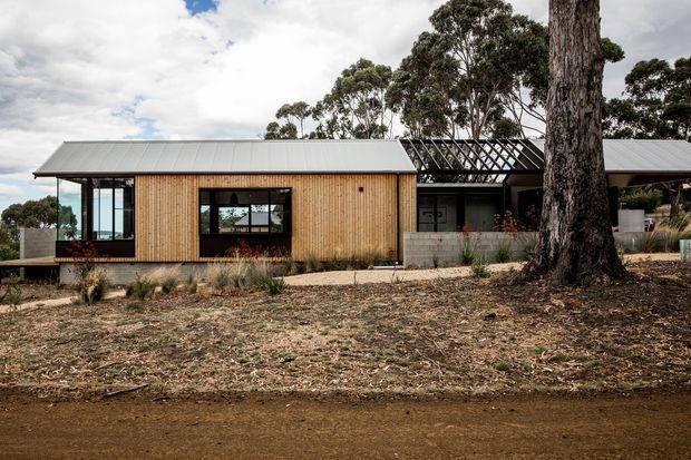 Single-storey contemporary residential building viewed from the front, featuring a long low-pitched metal roof, warm vertical timber cladding, extensive dark-framed glazing, and a central glazed courtyard or breezeway structure. The house sits in a dry, landscaped setting with mature eucalyptus trees and understated concrete masonry elements.
