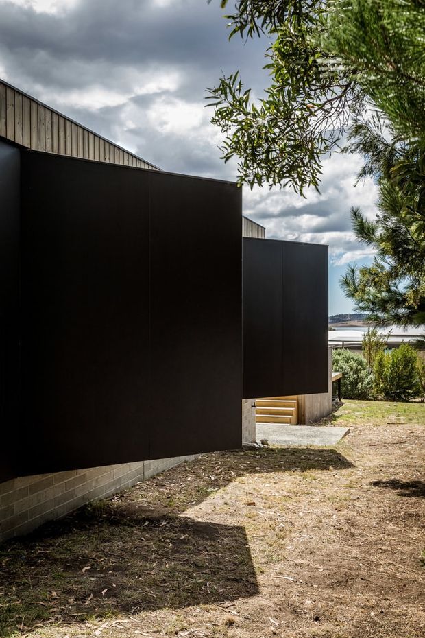 Exterior architectural view of a contemporary building with bold matte-black wall cladding, a projecting box-like volume, and a corrugated or vertically ribbed gabled roof form. The lower wall sits on a light grey concrete-block base, with timber steps leading toward the entrance. The building is set in a landscaped suburban site with trees and shrubs, and a body of water or distant landscape is visible beyond.
