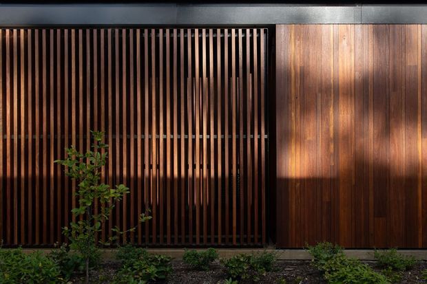 A close-up exterior view of a contemporary building facade featuring vertical dark timber battens screening a recessed opening alongside warm timber-clad wall panels. Low landscaping runs along the base, with strong sunlight and shadow creating a warm, textured composition.