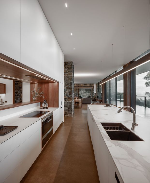 A long, refined contemporary kitchen with white full-height cabinetry, warm timber open shelving and wall panelling, dark stone feature columns, and a large white marble-look island with integrated double sink and curved tapware. The kitchen opens toward a dining and living area, with extensive floor-to-ceiling glazing along one side and linear pendant lighting overhead. The space has a warm, polished and highly functional feel.