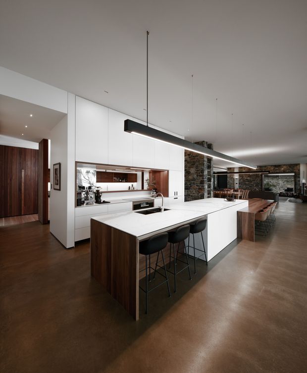 A spacious open-plan kitchen and dining area with a large white island, dark timber cabinetry, black bar stools, and a long linear pendant light. The kitchen opens to a dining and living area framed by exposed stone walls, with polished concrete flooring and a warm, contemporary material palette.