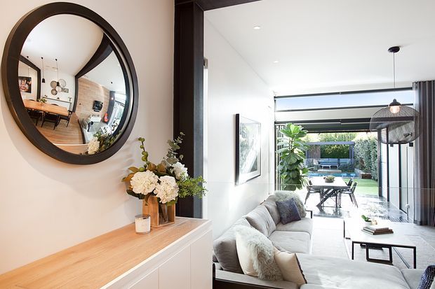 Bright contemporary open-plan living room viewed from an entry or circulation area, featuring a white sectional sofa, timber console cabinetry, dark timber-framed round mirror, coffee table, and full-height glazed opening to a landscaped outdoor dining area. The mirror reflects part of the adjacent dining and kitchen area. Dark structural framing and large glass openings create a strong indoor-outdoor connection.