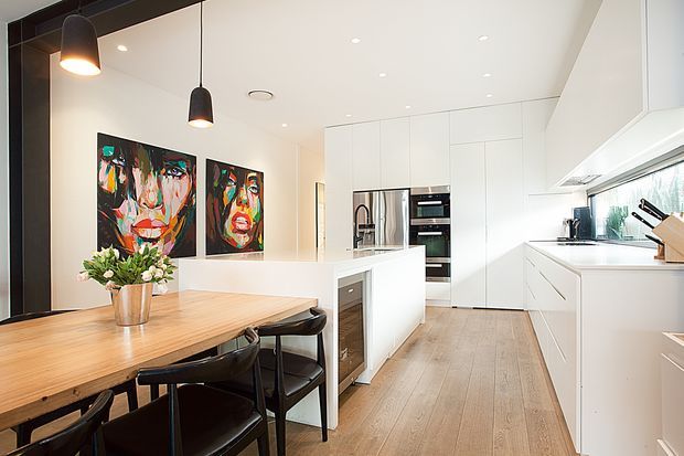 Bright contemporary open-plan kitchen and dining area with streamlined white cabinetry, a central island, integrated stainless-steel appliances, light timber flooring, and a substantial timber dining table with black chairs. Black pendant lights and colourful abstract artwork provide contrast against the predominantly white fit-out.