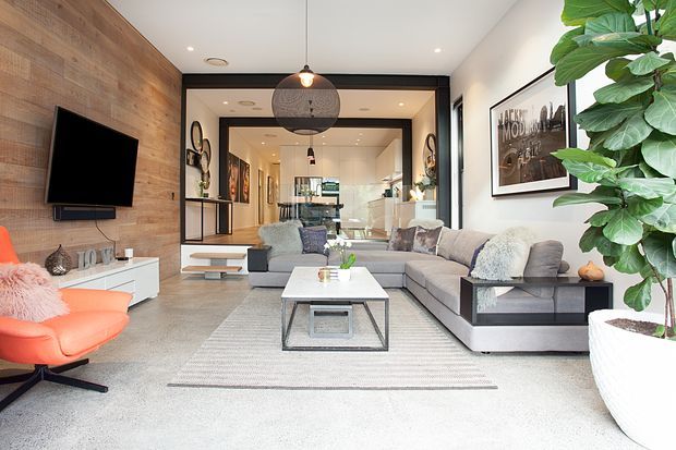 Bright contemporary living room with a large grey sectional sofa, white rectangular coffee table, textured area rug, and a vivid coral lounge chair. A warm timber feature wall with wall-mounted television anchors the room, while black-framed glazed partitions open toward a rear kitchen or dining area. Pendant lighting, artwork, shelving, and a large indoor fiddle-leaf plant complete the space.