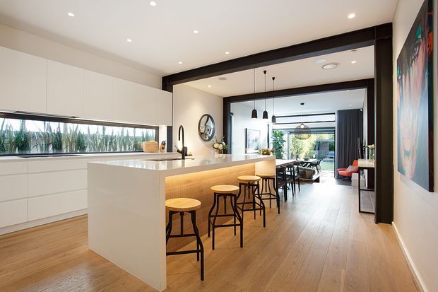 Bright open-plan contemporary kitchen with a large white island, integrated cabinetry, timber flooring, and four black-framed timber stools. The kitchen opens directly into a dining and living area through a wide opening framed by a dark structural beam, with extensive glazing and garden views beyond. Recessed ceiling lights, pendant lights, and a dark artwork add contrast to the light, warm-toned interior.